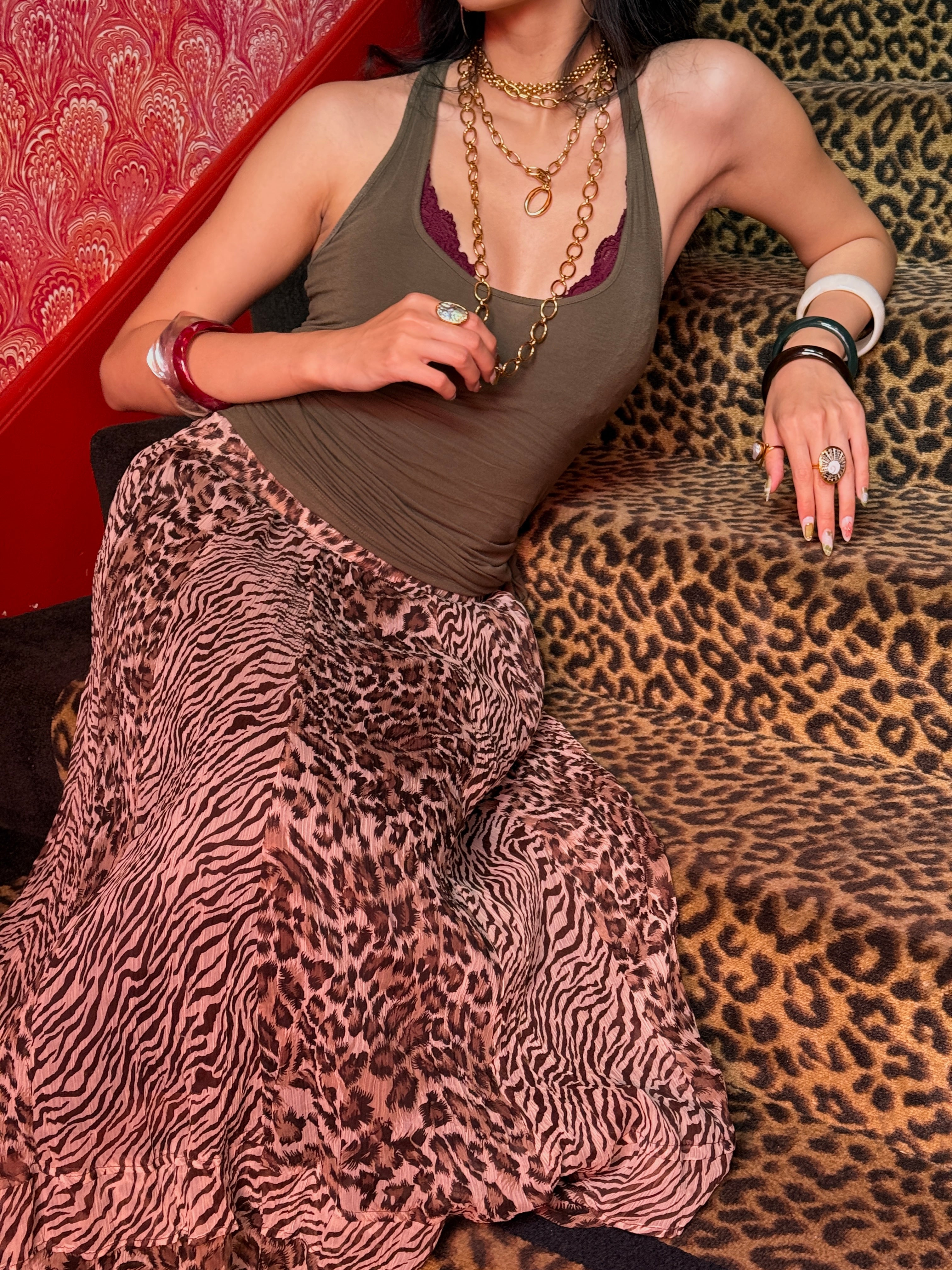 A woman reclining on a leopard-print sofa, wearing a sleeveless olive-brown top and a long pink leopard-print skirt. She is accessorized with layered gold necklaces, rings, and bracelets.